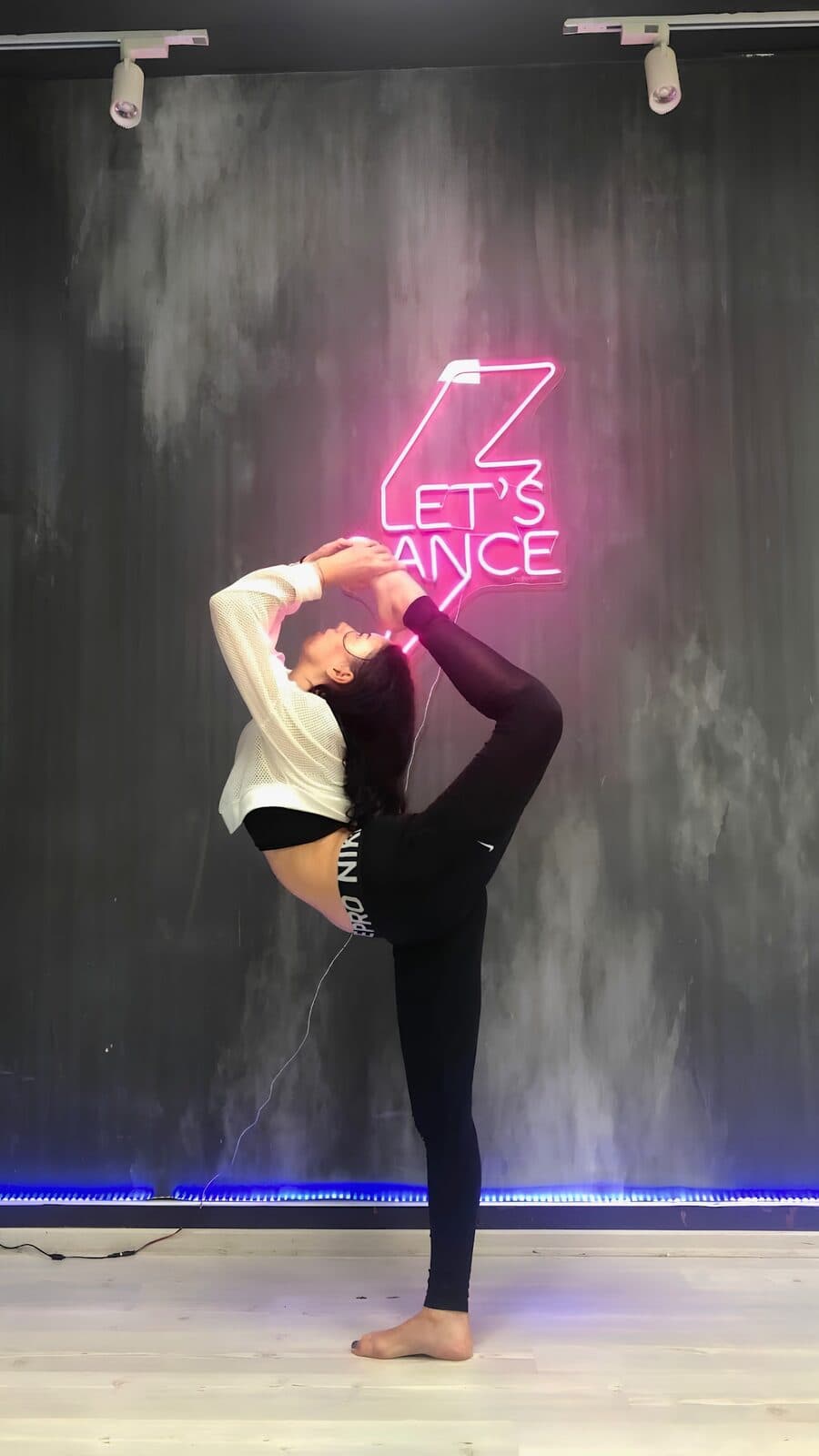Tuğba'Danspor stüdyosunda "Let's Dance" neon yazısının önünde esneklik hareketi yapan dansçı